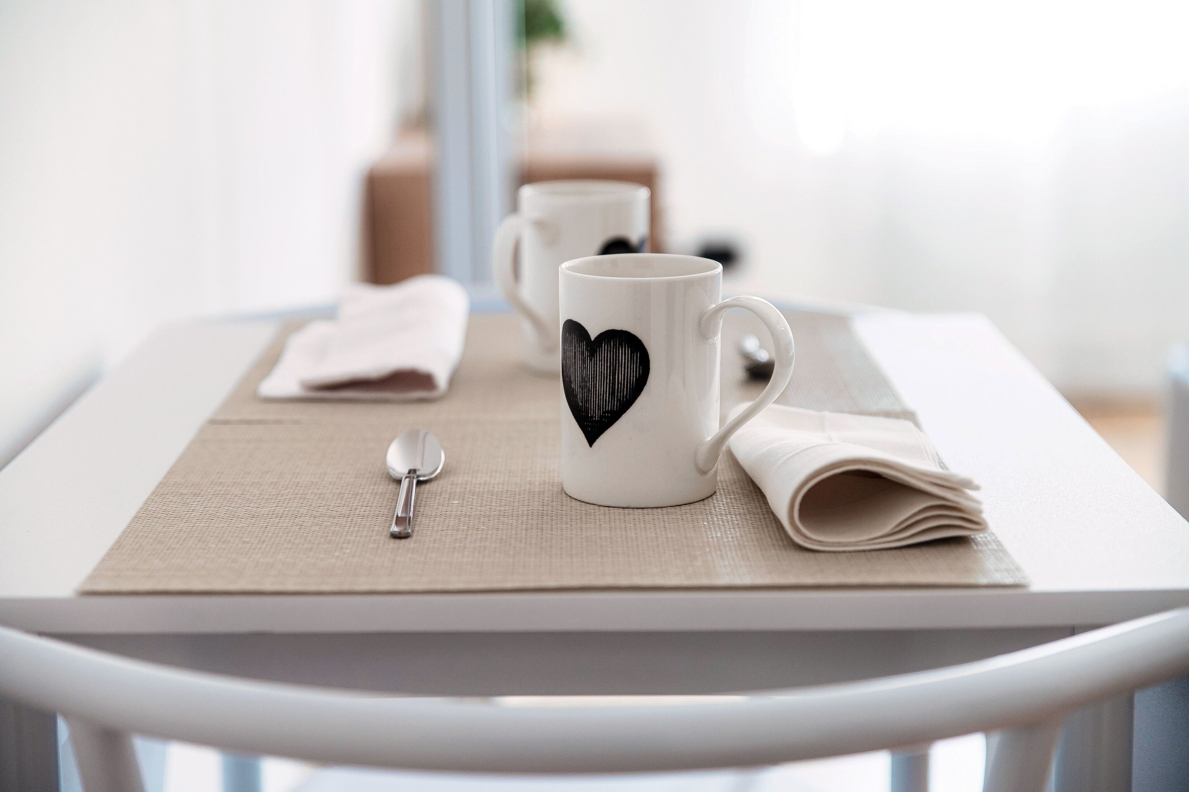 A table with two white mugs, one with a black heart design, a folded beige napkin, and a spoon on a beige placemat.