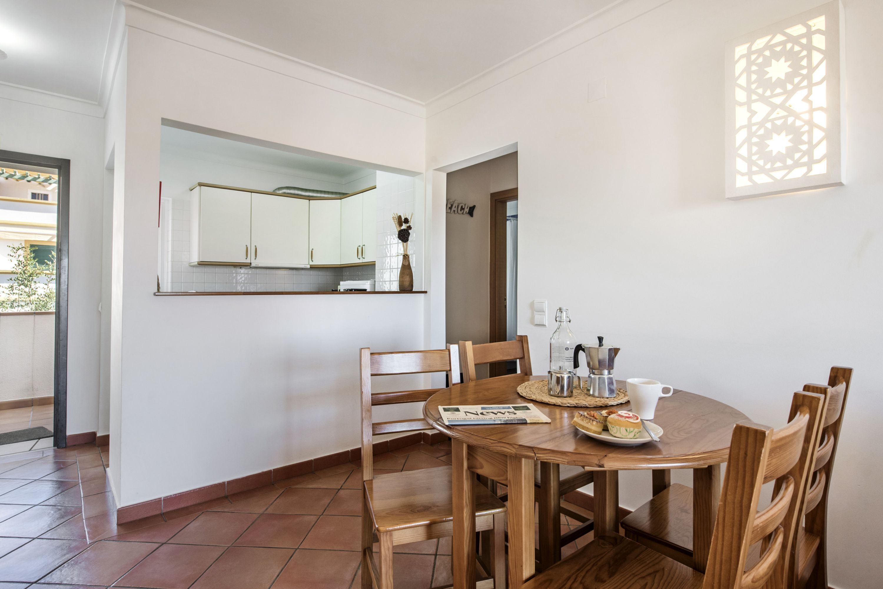 A dining area with a round wooden table, six chairs, a small kitchen in the background, and a decorative wall piece.