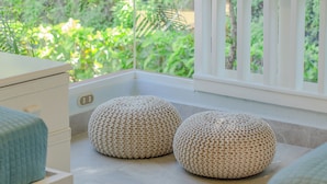 A room with a large window, a bed, two round poufs, and a wooden nightstand.