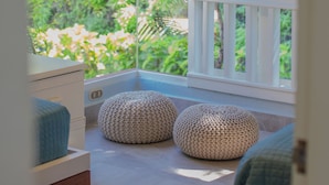 A room with a view of greenery through a window, a ceiling fan, and two round ottomans.