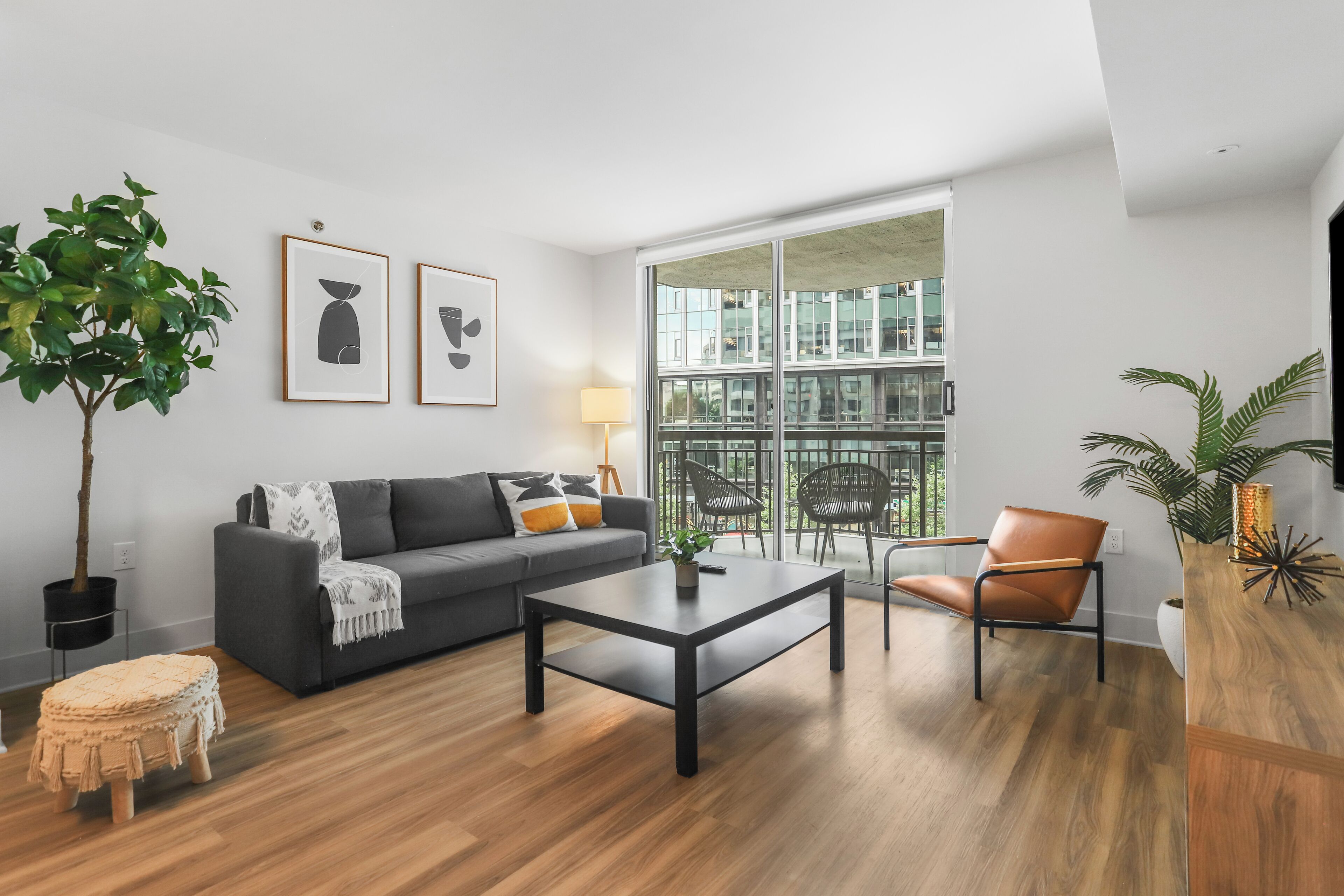 Un salon moderne avec un canapé gris, une table basse noire et un plancher en bois. Une grande fenêtre offre une vue sur les bâtiments, et une plante est placée sur le côté gauche.