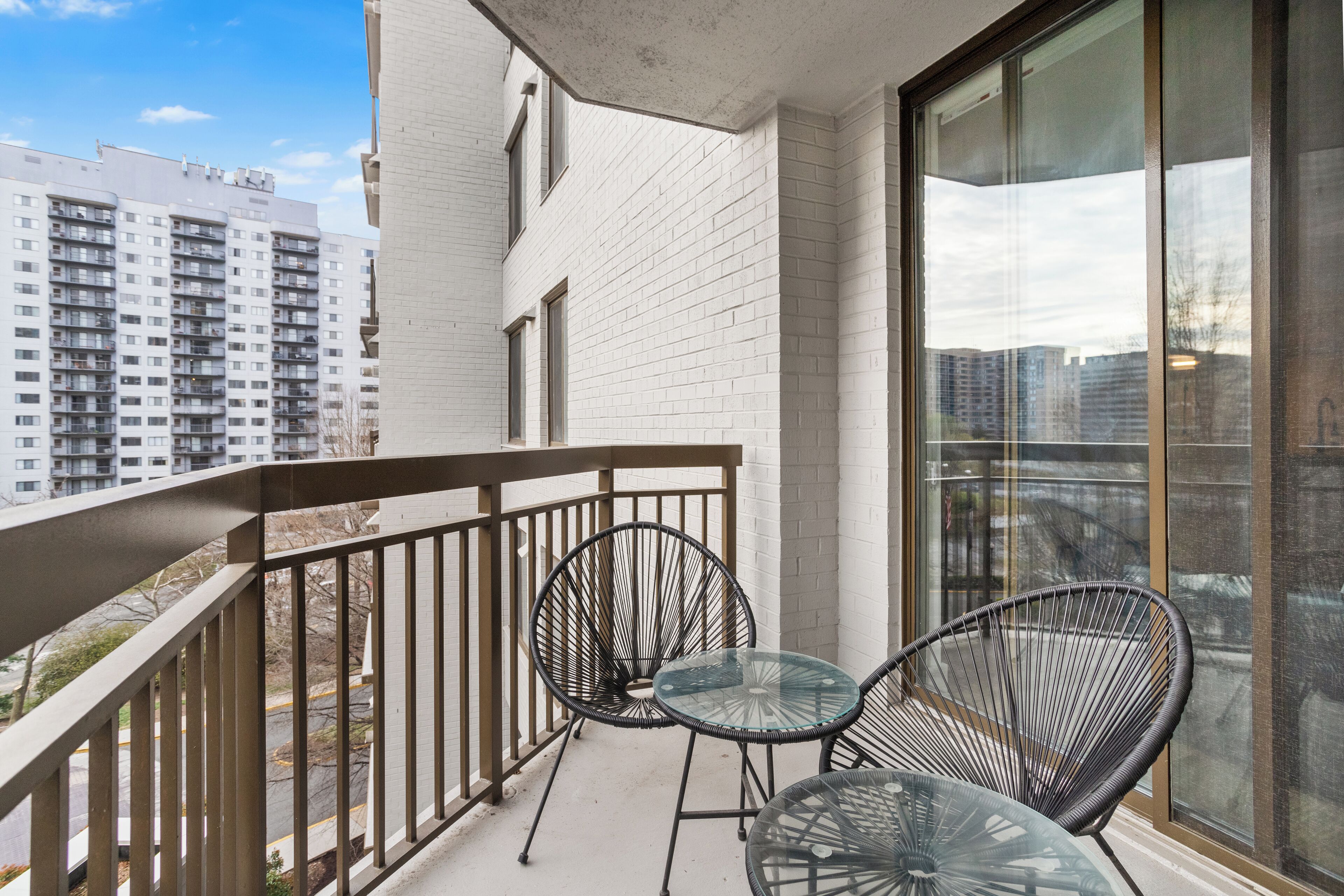 Un balcon avec deux chaises en fil de fer noir et une table en verre, d’où l’on peut admirer un panorama urbain avec de hauts immeubles.