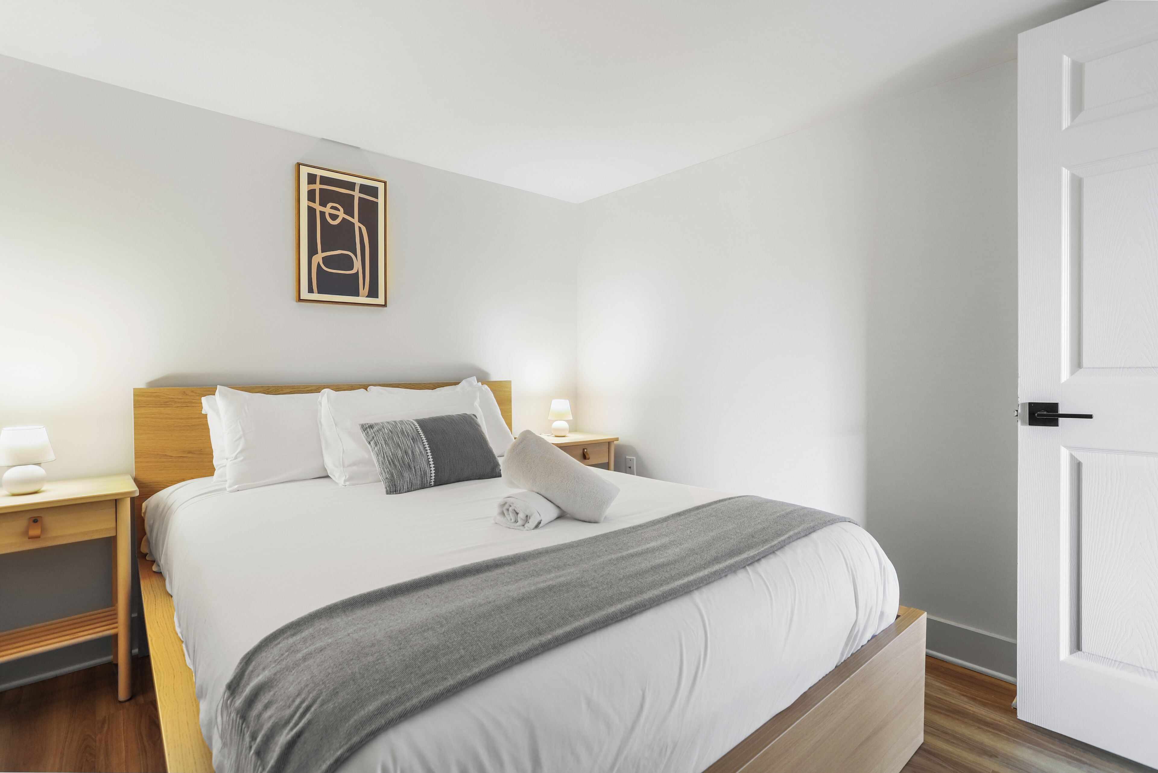 Una cama bien hecha con una manta gris, almohadas blancas y un cabecero de madera. Hay una mesita de noche con una lámpara y un pequeño objeto decorativo. Encima de la cama, en la pared, cuelga un cuadro enmarcado.