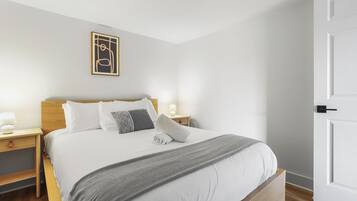 Una cama bien hecha con una manta gris, almohadas blancas y un cabecero de madera. Hay una mesita de noche con una lámpara y un pequeño objeto decorativo. Encima de la cama, en la pared, cuelga un cuadro enmarcado.