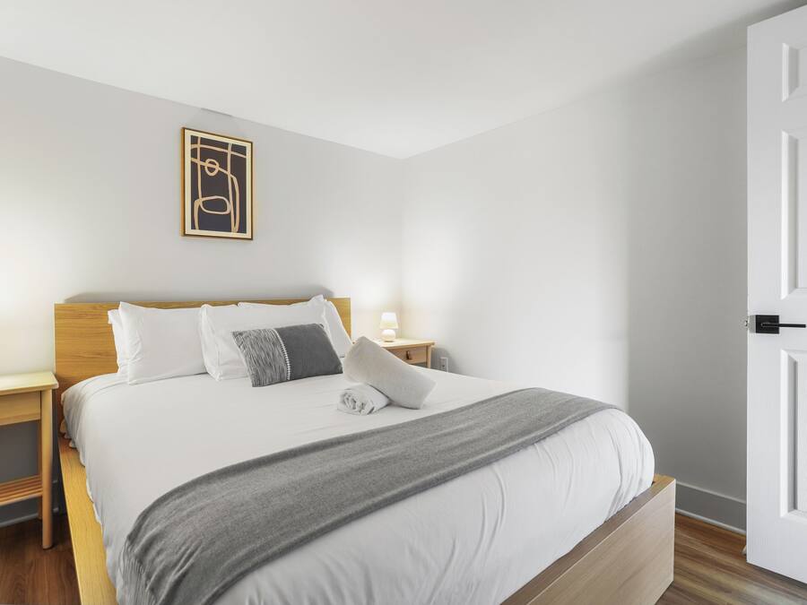Una cama bien hecha con una manta gris, almohadas blancas y un cabecero de madera. Hay una mesita de noche con una lámpara y un pequeño objeto decorativo. Encima de la cama, en la pared, cuelga un cuadro enmarcado.