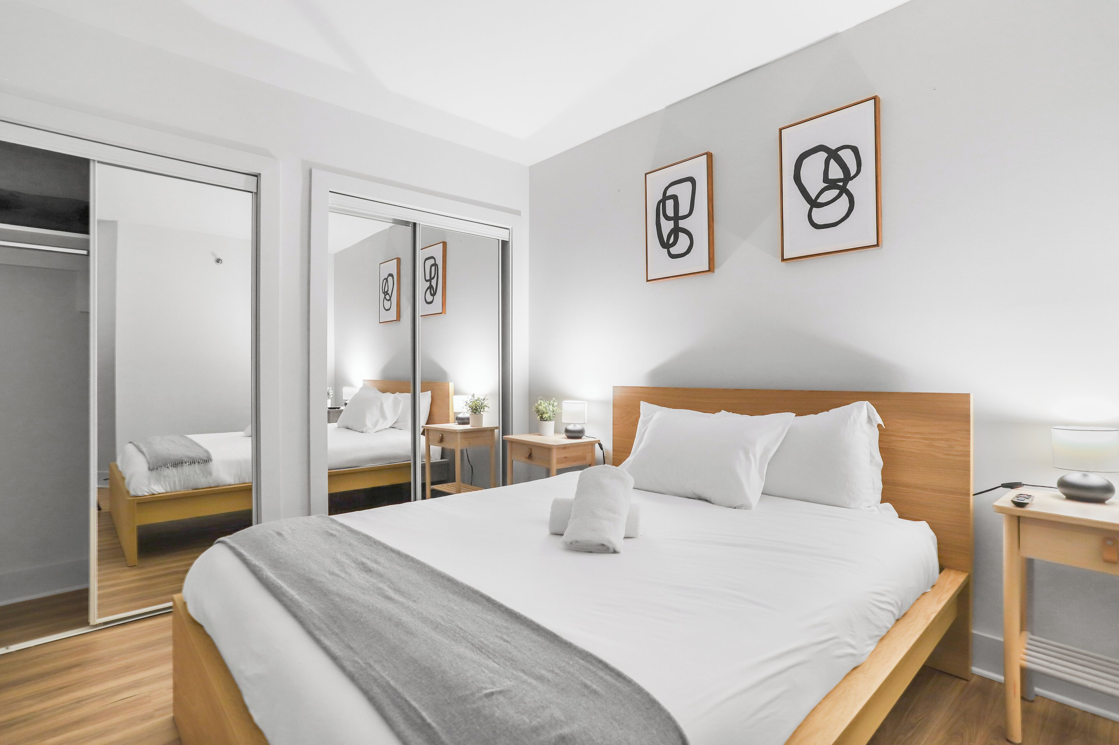 A modern hotel room with a large bed, a wooden headboard, a bedside table with a lamp, and a mirror. There are framed abstract artworks on the wall.