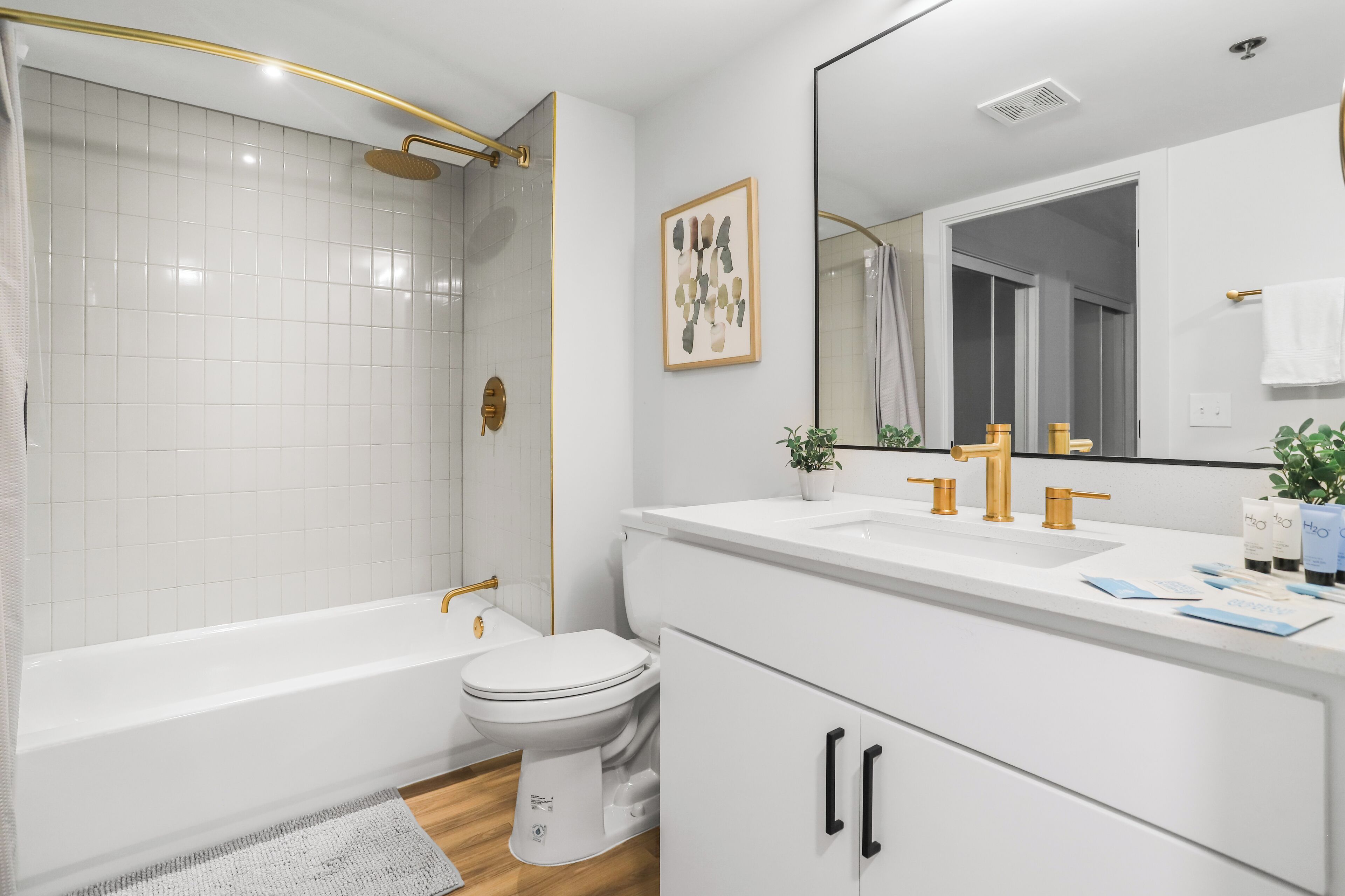 A modern bathroom with a white bathtub, toilet, and sink. There is a shower with a gold showerhead and a framed artwork on the wall.