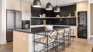 Una cocina moderna con una isla central, taburetes de bar, iluminación colgante y electrodomésticos de acero inoxidable.