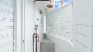 A narrow hallway with white walls, a tiled floor, and a white door on the left. There is a shower area with a showerhead and pipes on the right.