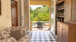A room with a checkered floor, a sofa with patterned cushions, a dining table with chairs, and a view of a lush garden through open shutters.