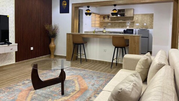 A modern living room with a sofa, a glass coffee table, and a kitchen area with a bar. - Yellow Door Aparment (Abuja)