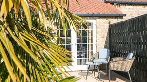 A patio area with a stone wall, a wicker chair, a table, and a glass door leading to a room with a tiled roof. - Rose Cottage (Nottingham)