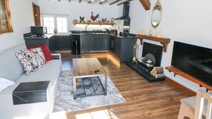A modern living room with a sofa, coffee table, and fireplace. A kitchen with a chimney and rooster decorations is visible in the background. - Rose Cottage (Nottingham)