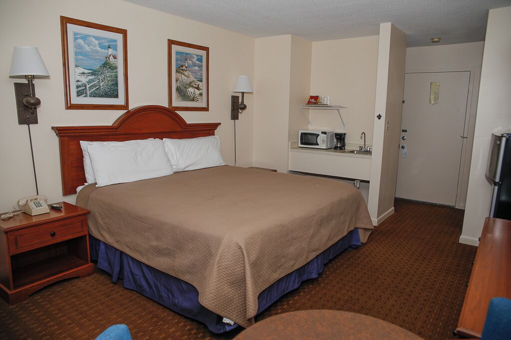 A hotel room with a large bed, a bedside table with a lamp, a microwave, and a small shelf with toiletries.