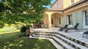A patio with outdoor seating, a staircase leading to a covered area, and a building with a tiled roof and green shutters.