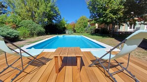 A wooden deck with a pool, surrounded by greenery and a house in the background.