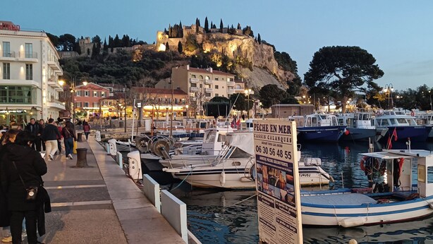 A marina with boats docked, a castle on a hill in the background, and people walking along the waterfront.
