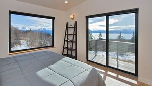 Ein Schlafzimmer mit einem großen Bett, zwei Fenstern mit Blick auf Berge und See, einer Leiter und einem kleinen Regal.