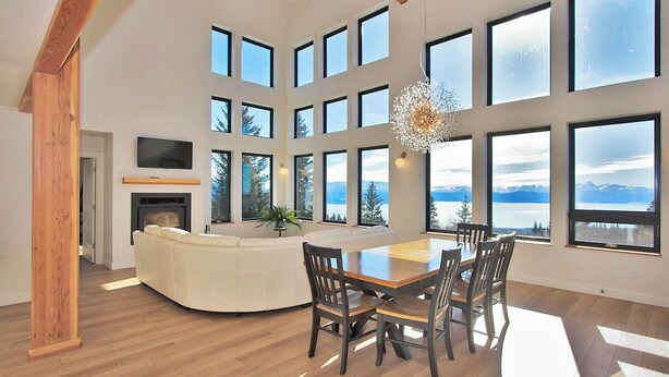 Ein geräumiges Wohnzimmer mit einer großen Holzbalkendecke, einem Kamin, einem weißen Sofa, einem Esstisch mit Stühlen und großen Fenstern mit Blick auf Berge und einen See.