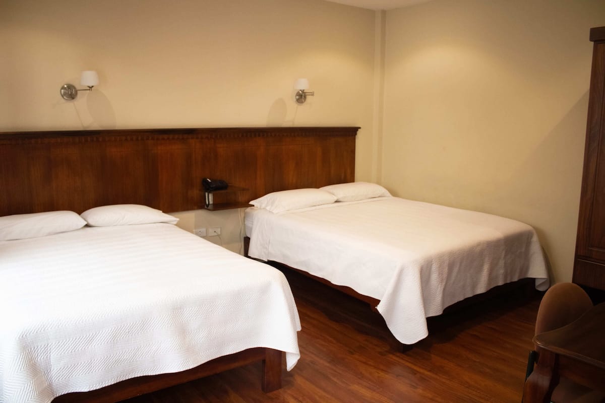 A hotel room with two single beds, wooden headboard, bedside tables, and wall-mounted lamps.