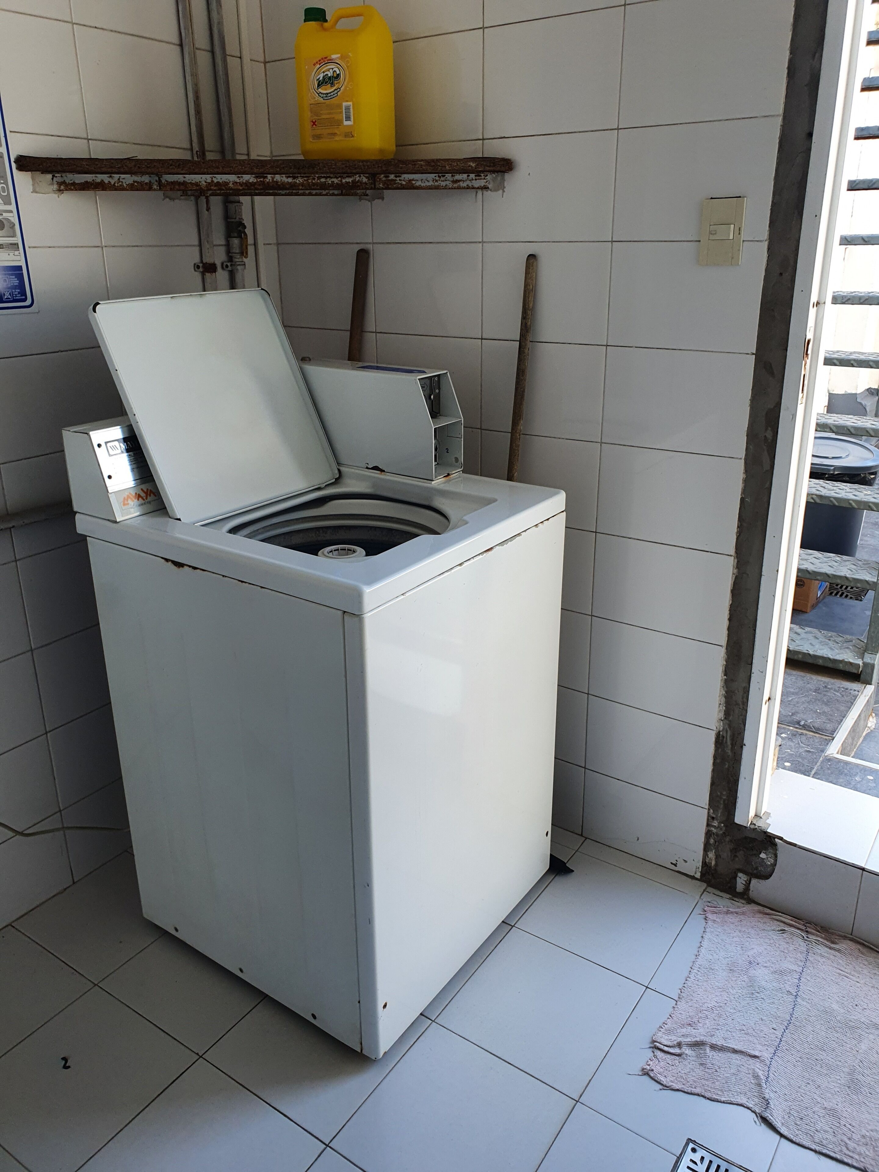 Eine kompakte, weiße Waschmaschine in einer kleinen Waschküche mit gefliesten Wänden und einer sichtbaren Tür, die nach draußen führt.