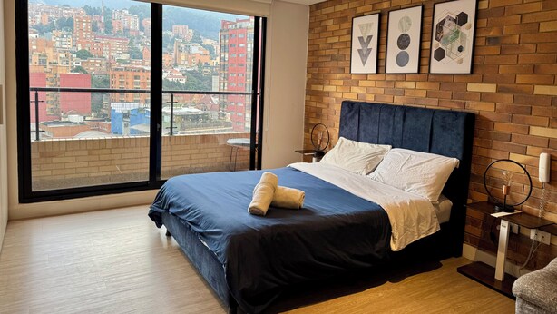A modern bedroom with a large window, a brick accent wall, and a bed with a blue headboard.