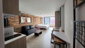A modern hotel room with a kitchen area, a living room with a sofa and coffee table, a large bed, and a dining area.