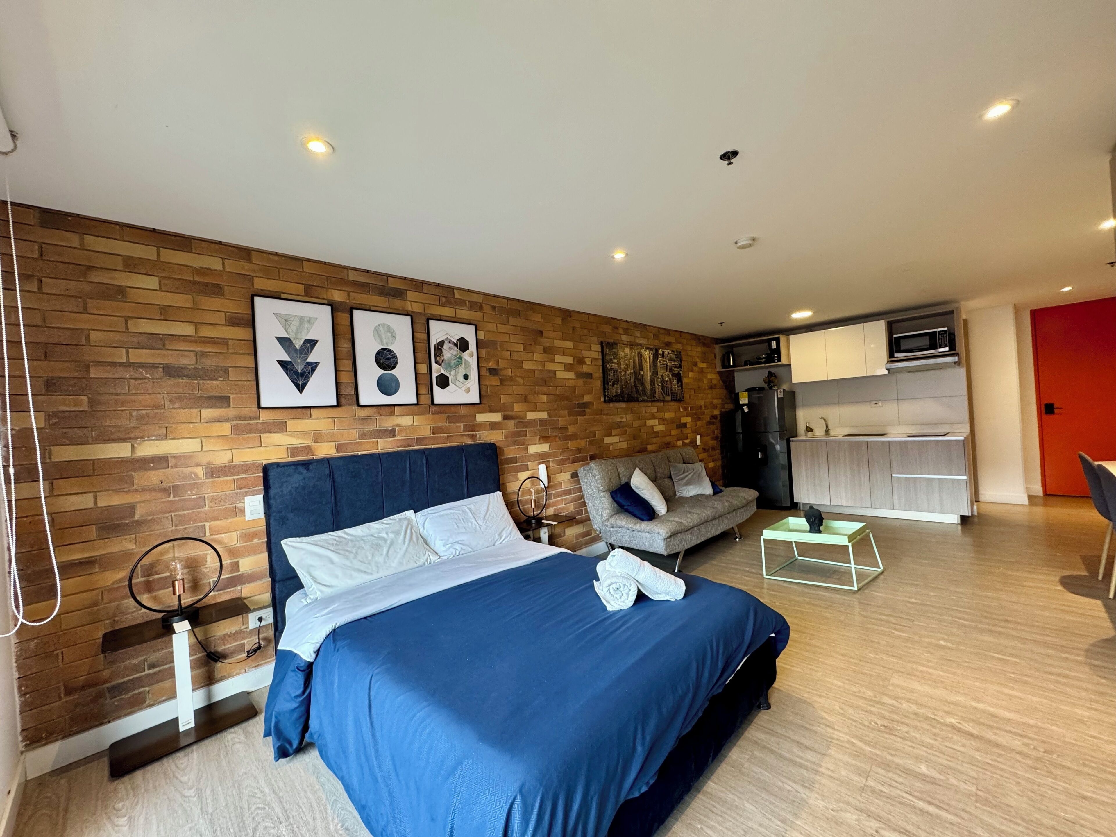 Habitación de hotel moderna con pared de ladrillo, una cama grande con colcha azul, un sofá gris, una mesita pequeña y una zona de kitchenette.