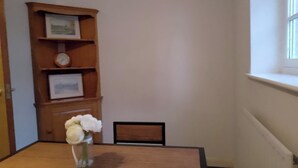Ein Holztisch mit einer Vase voller weißer Blumen, ein Holzregal mit gerahmten Bildern und einer Uhr sowie ein Fenster mit Jalousien.