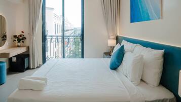 Ein Hotelzimmer mit einem großen Bett, einem Schreibtisch mit Spiegel, einem kleinen Tisch mit Lampe und Blick auf die Stadt vom Balkon aus.