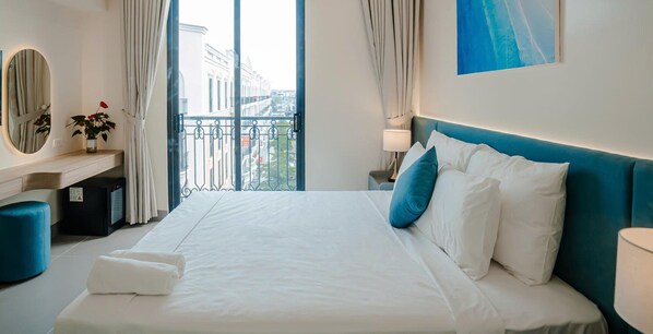 A hotel room with a large bed, a desk with a mirror, a small table with a lamp, and a view of the city through a balcony. - Lotus Sam Son Hotel (Sam Son)