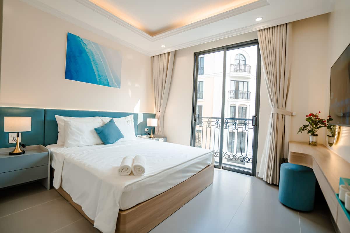 A modern hotel room with a large bed, a nightstand, a bedside lamp, a blue accent wall, a view of a balcony, and a painting on the wall.