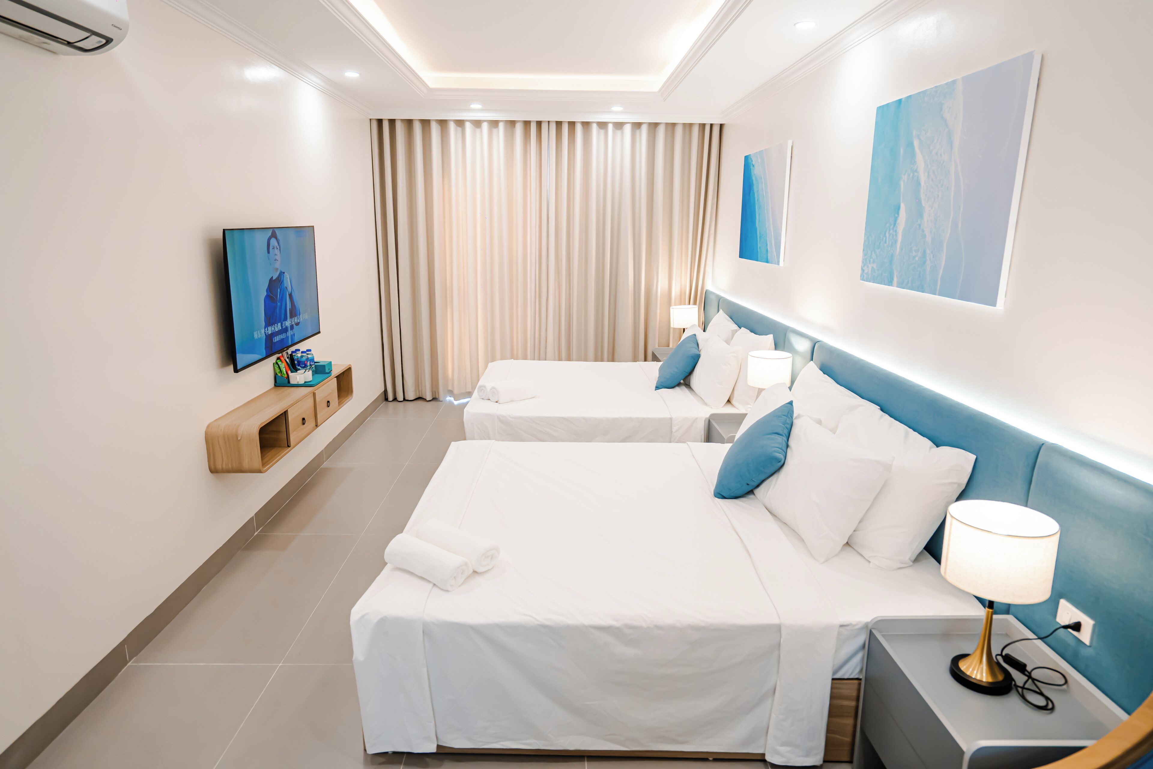 A hotel room with two beds, a TV, a desk, and a window with curtains.