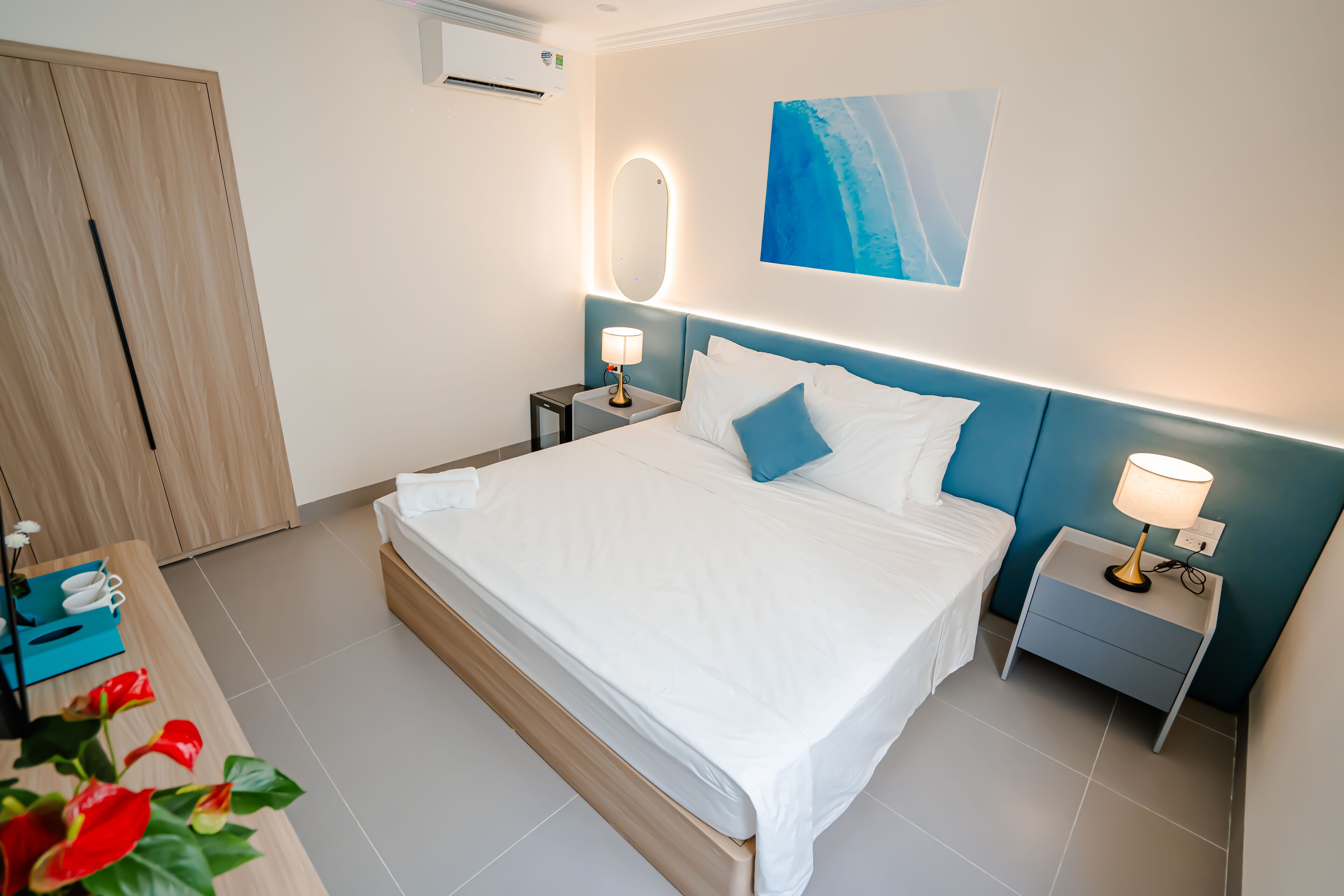A modern hotel room with a large bed, bedside tables with lamps, a wooden wardrobe, and a blue abstract painting on the wall.