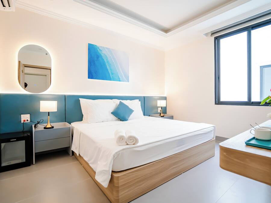 A hotel room with a bed, bedside tables, a mirror, a painting, a window, and a desk with a cup and a plant.