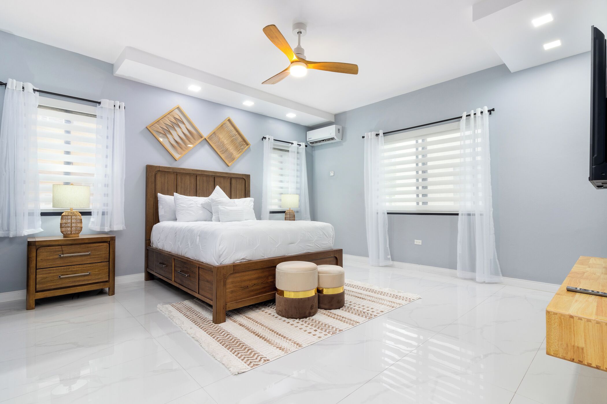 Une chambre moderne avec un lit en bois, un ventilateur de plafond et une grande fenêtre ornée de rideaux blancs.