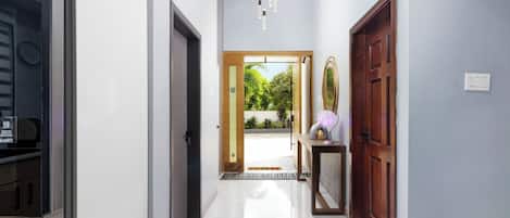A hallway with a modern kitchen area on the left, a wooden door on the right, and a view of greenery outside.