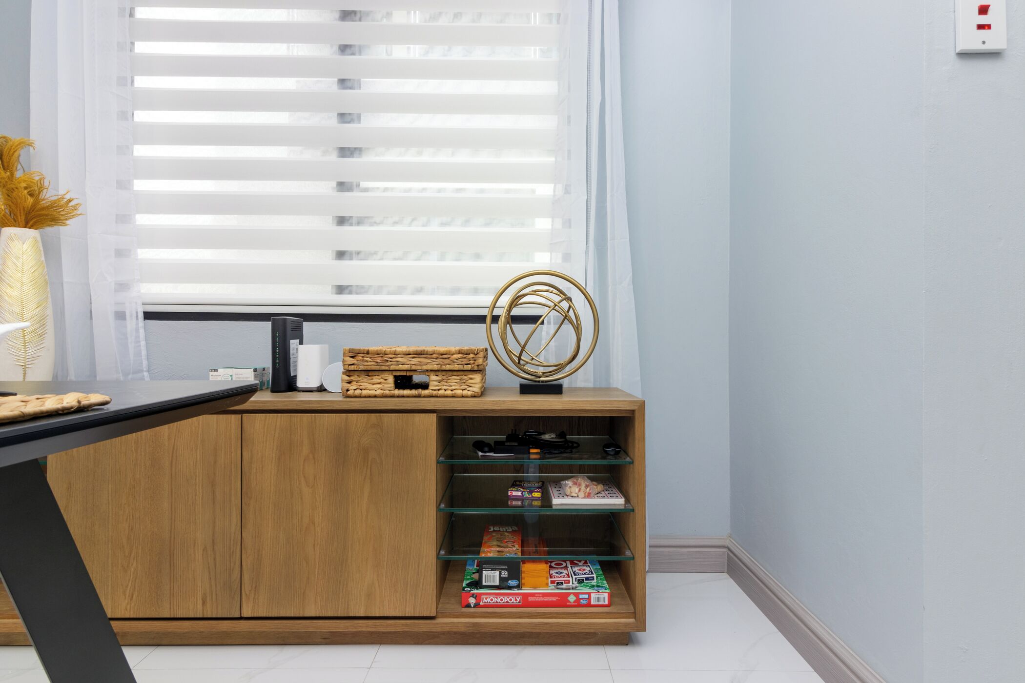 Een kamer met een houten bureau, een raam met zonwering, een decoratieve gouden ring, en planken met gezelschapsspellen.
