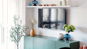A modern living room with a flat-screen TV mounted on the wall, a green cabinet below, and a variety of decorative items on a shelf above. - Como Hills (Como)