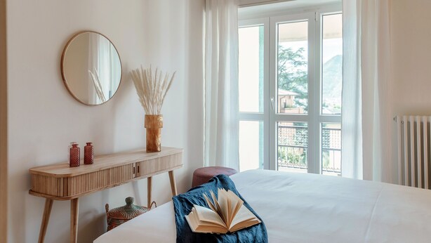 A hotel room with a bed, a bedside table with a mirror, a window with curtains, and a view of the outside. - Como Hills (Como)