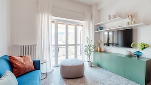 A modern living room with a blue sofa, a round ottoman, a green cabinet, and a large window with white curtains. - Como Hills (Como)