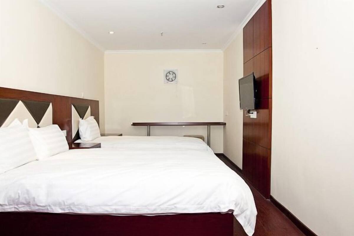 A hotel room with a large bed, a bedside table, a flat-screen TV mounted on the wall, and a clock on the wall.