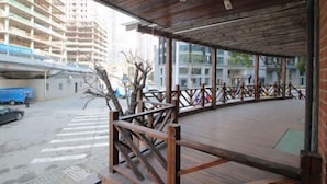 A covered outdoor area with wooden flooring and railings, overlooking a street with buildings and a blue truck.
