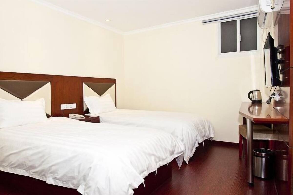 A hotel room with two single beds, a wooden headboard, a small table with a kettle, and a trash bin.