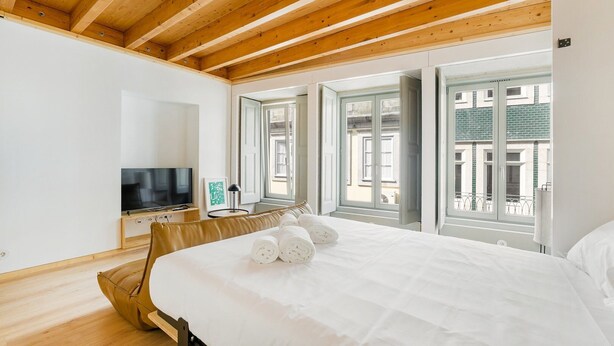 A bedroom with a wooden ceiling, a bed with white linens, a TV on a wooden stand, and large windows with a view of buildings.