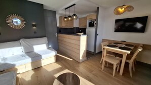 En kompakt opholdsstue med sofa, spisebord med stole, et køkken og et fjernsyn ophængt på væggen.