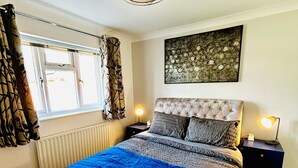 A bedroom with a tufted headboard, a large bed with a blue blanket, a hanging light fixture, a window with curtains, and a wall-mounted abstract art piece. - Beautiful, fully equipped, pet friendly 3 bedroom house in Shakespeare County. (Alcester)