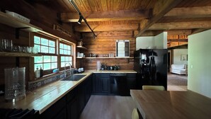 A rustic kitchen with wooden beams, a black refrigerator, and a window overlooking greenery. - Beautiful Log House  4 Bed, 2 Baths, 15 acre land, close to Madison, sleeps 13 (Albany)
