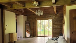 A spacious room with a wooden ceiling, a chandelier, a sofa with patterned cushions, and a large window offering a view of the outdoors. - Beautiful Log House  4 Bed, 2 Baths, 15 acre land, close to Madison, sleeps 13 (Albany)