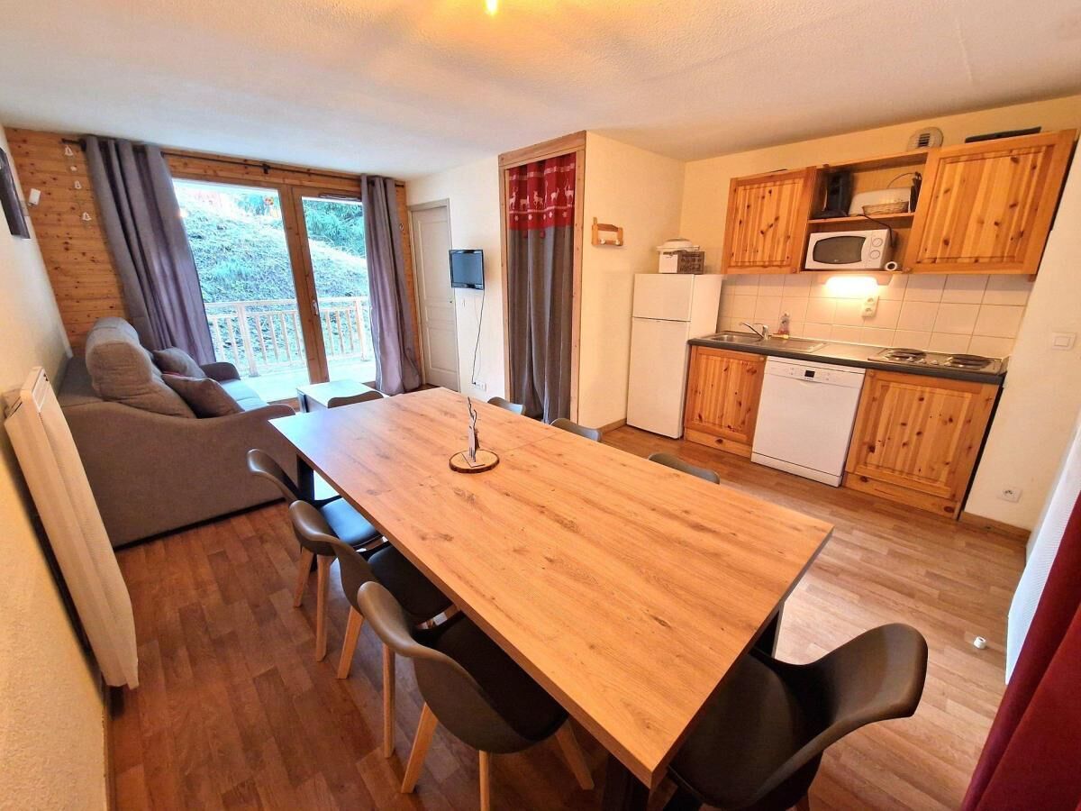 Un espace de vie compact comprenant une table à manger, des chaises, un canapé, un coin cuisine avec des meubles en bois et des appareils électroménagers, ainsi qu’une porte-fenêtre coulissante donnant sur un espace extérieur.